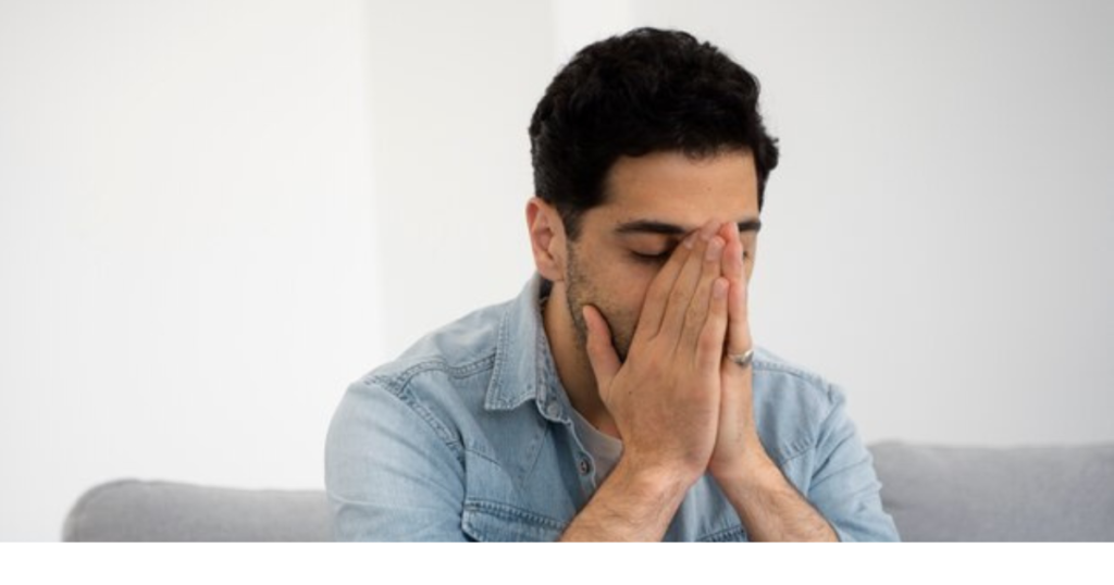 A man in a denim shirt sitting on a couch with his face in his hands, expressing stress or sadness.